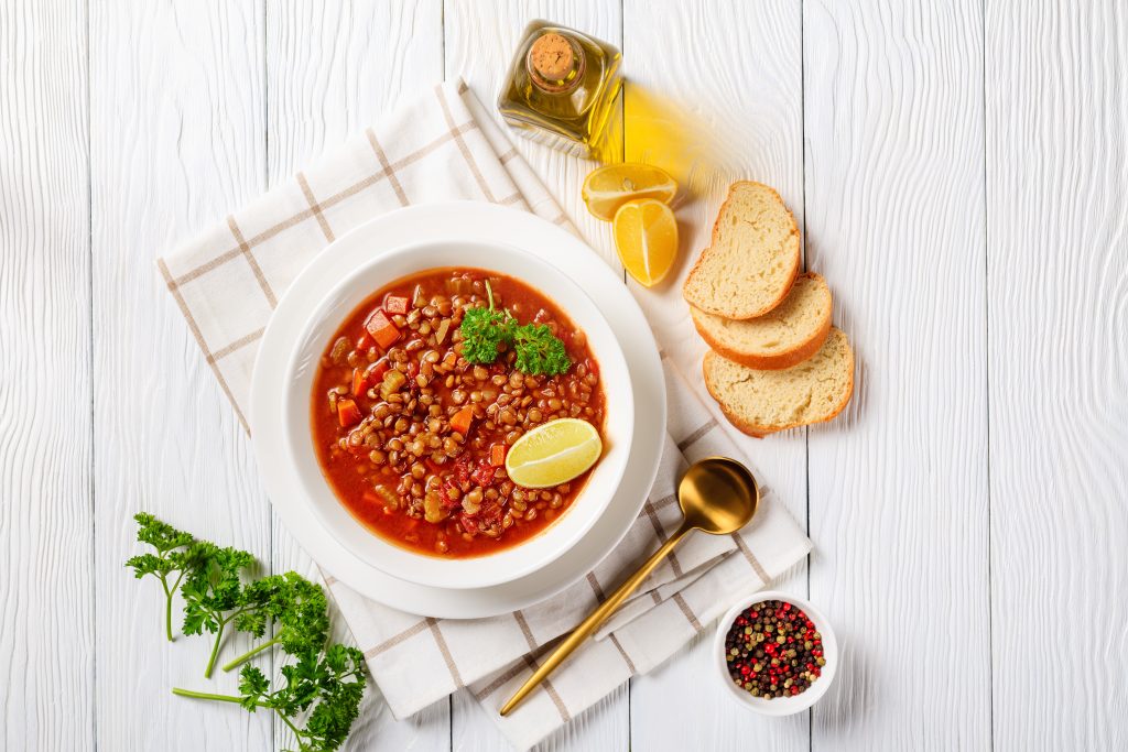 Curried Lentil Chickpea Stew, Lentil soup with vegetables and lemon wedges in a white bowl on a wooden table with a golden spoon and toasts, horizontal view from above, flatlay, free space